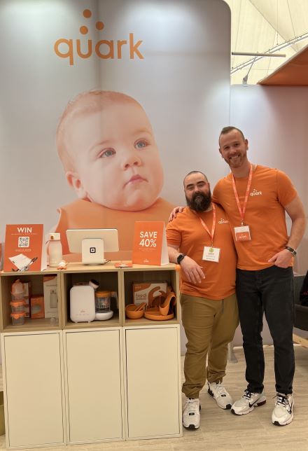 Quark 联合创始人 Senez 和 Gurinskas 在展会的展位站在他们的产品旁边。背后有婴儿照片和他们的标志。
